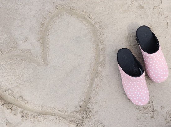 Roze klompen met witte stippen op het strand naast een hart in het zand.
