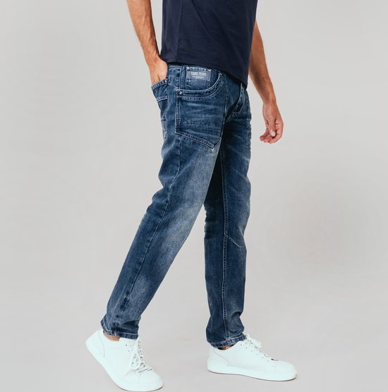 Man in een donkerblauwe trui en een stonewashed blauwe jeans. De jeans heeft extra naden op de bovenbenen en witte sneakers.