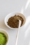 Wit schaaltje met hojicha poeder en een bamboe schepje, naast een schaaltje met matcha.