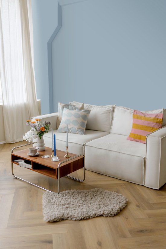 Een woonkamer met een poederblauwe muur, een witte bank met gekleurde kussens, een salontafel met bloemen en kaarsen.
