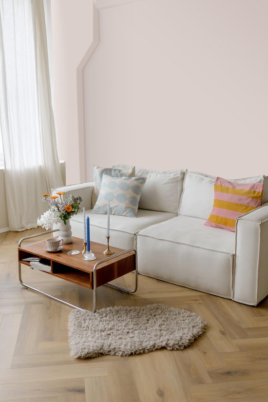 Een lichte woonkamer met een witte stoffen hoekbank, een houten salontafel met metalen frame en decoraties, en een lichtroze muur.
