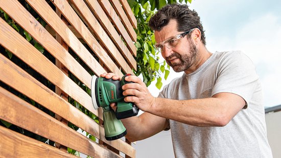 Man met veiligheidsbril schuurt een houten schutting met groene Bosch excenterschuurmachine.
