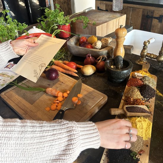 Een keukenblad met ingrediënten, een hand die een recept vasthoudt en kruiden uitgespreid op een snijplank.