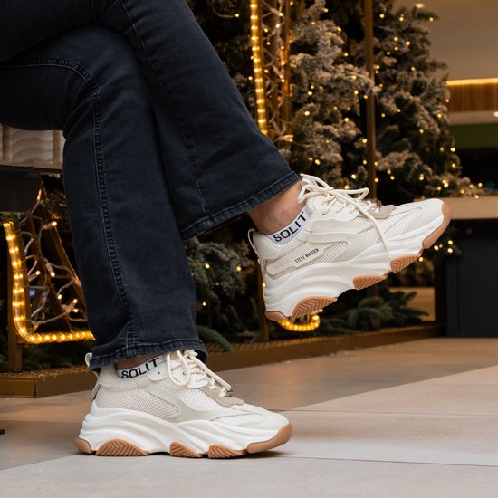 Crème lage sneakers met dikke witte zolen en bruine accenten, gedragen met een spijkerbroek. De sokken hebben de tekst "SOLIT".