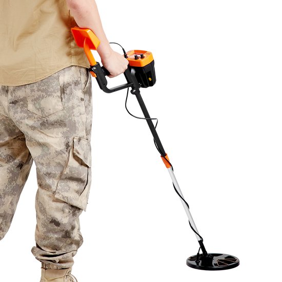 Man in camouflagebroek en bruin shirt met een oranje metaaldetector, gefotografeerd tegen een witte achtergrond.