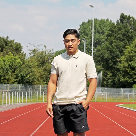 Man op een atletiekbaan in een beige polo en zwarte sportshort.