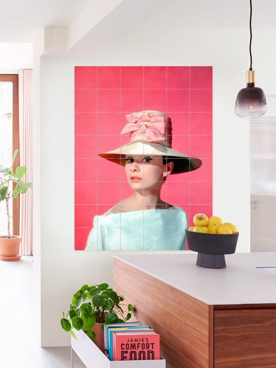 Audrey Hepburn portant un grand chapeau, un nœud rose et un haut pâle, sur fond rose, dans une cuisine avec comptoir et buffet en bois.