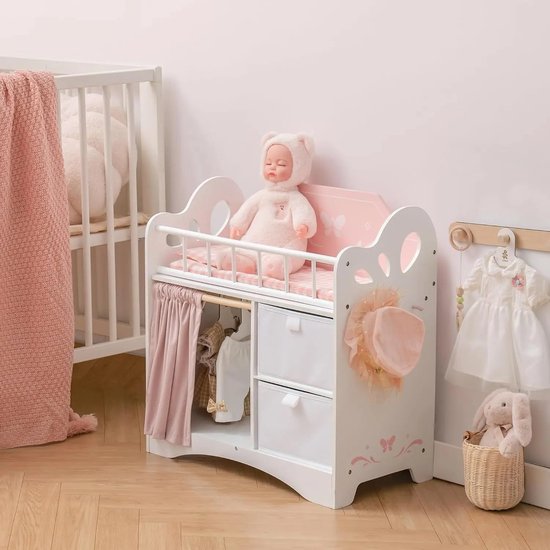 Wit poppenbedje met roze beddengoed, vlinderdecoraties, roze gordijn en opbergvakken. Poppenkleding hangt aan haken.