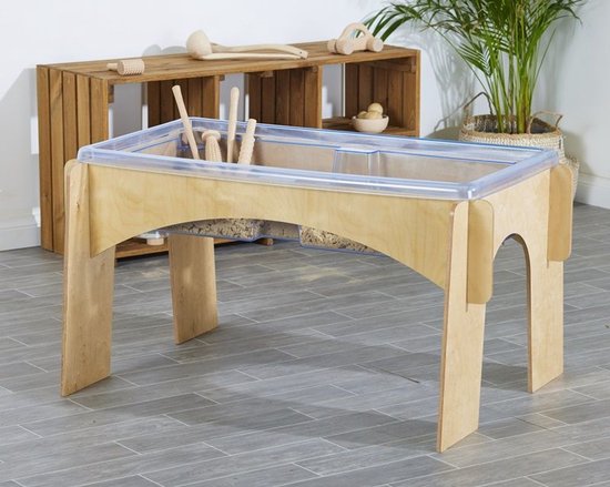 Houten zand- en watertafel gevuld met zand en speelgoed, met planten op de achtergrond.