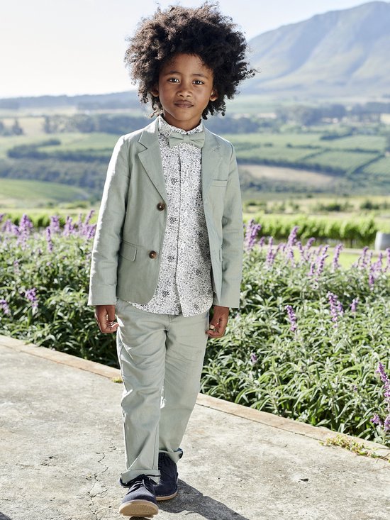 Jongen in lichtgroen pak: colbert, broek, wit printblouse en strikje. Buiten, met paarse bloemen.