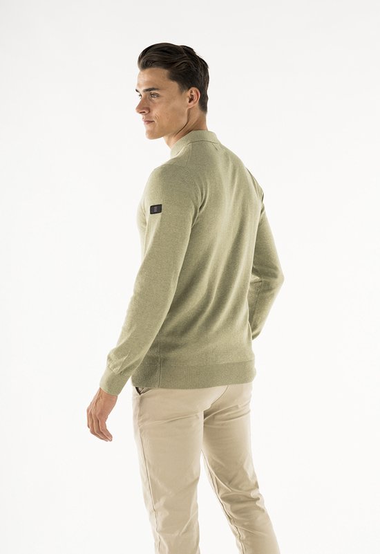 Man kijkt opzij, draagt een olijfgroene gebreide polo met lange mouwen en een label op de mouw.