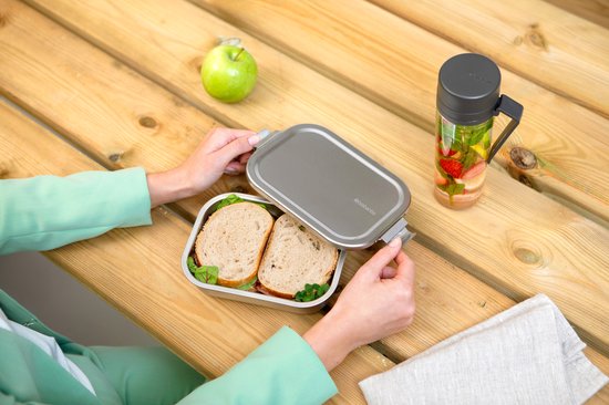 Personne en tailleur vert clair ouvrant une boîte à lunch en inox à une table en bois.