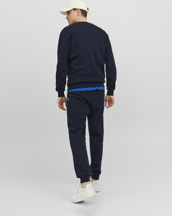 Man, achterkant, in donkerblauwe sweater en joggingbroek, met een beige pet en witte sneakers.