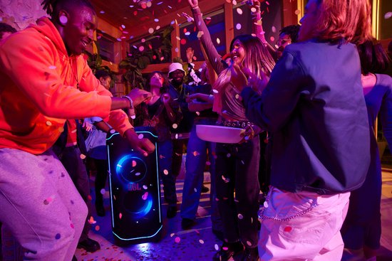 Zwarte JBL PartyBox Ultimate speaker met blauwe LED-verlichting, midden in een feestje met confetti en lachende mensen.