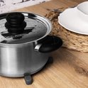 Een donkergrijze metalen pan met zwarte deksel en handvatten staat op een houten tafel naast een wit bord.