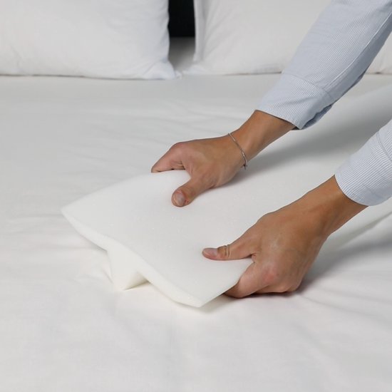 Handen houden een witte, schuimrubberen matraswig vast op een wit bed met witte lakens.