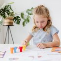 Jong meisje met blond haar in paardenstaart, gekleed in een blauw gestreept shirt met bloemen, dat aan een tafel aan het knutselen is met stempels.