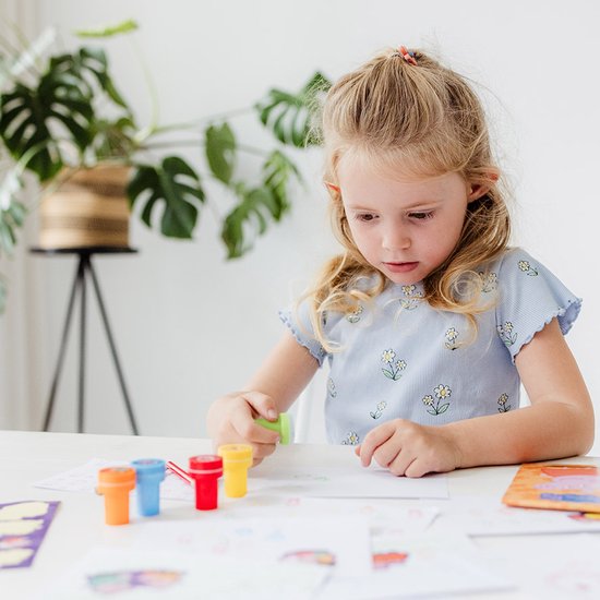 Jong meisje met blond haar in paardenstaart, gekleed in een blauw gestreept shirt met bloemen, dat aan een tafel aan het knutselen is met stempels.