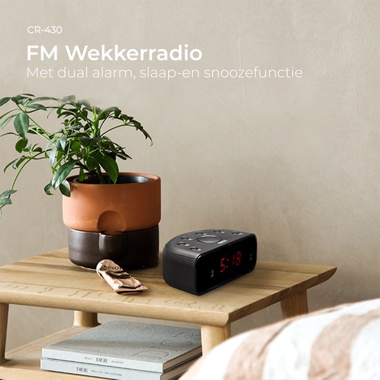 Zwarte wekkerradio met rode digitale tijdsaanduiding 5:18 op een nachtkastje naast een plant.
