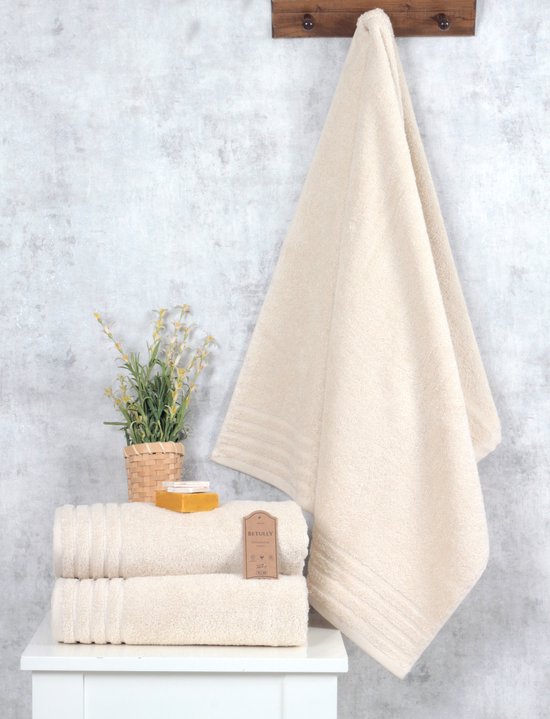 Een stapel van drie zachte, crèmekleurige handdoeken op een witte tafel. Een grote, crèmekleurige handdoek hangt ernaast.