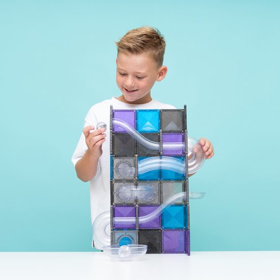 Een jongen met een witte T-shirt houdt een knikker vast en kijkt naar een Connetix knikkerbaan van paarse, blauwe en grijze tegels.