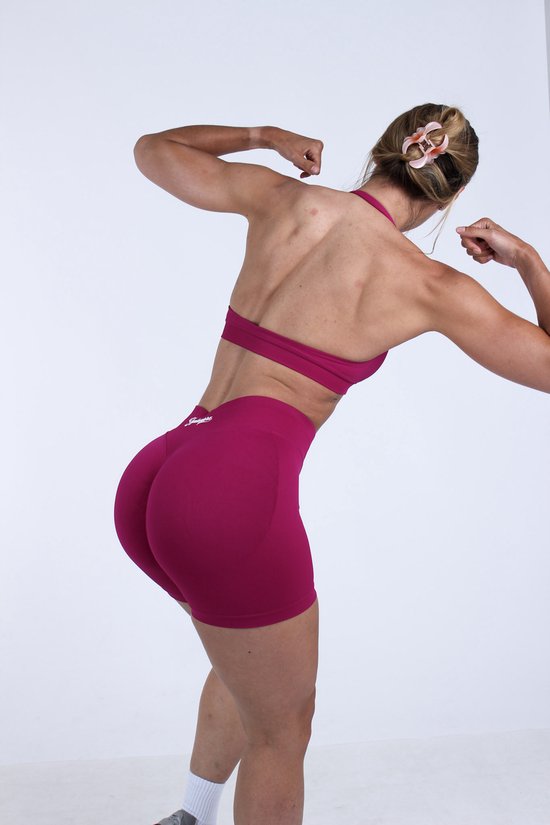 Vrouw in felroze sportbeha en short, rug naar de camera, die haar spieren toont.