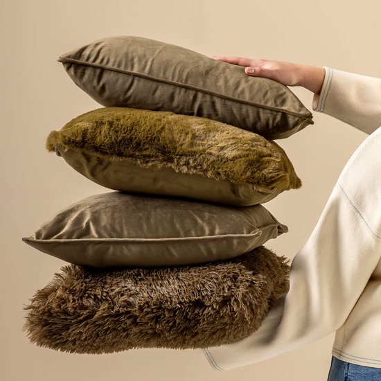 Een vrouw in een gebreide trui met blauwe stiksels houdt een stapel van vier kussens vast; twee van fluweel in olijfgroene tinten en twee met een lange, harige textuur in donkerbruine tinten.