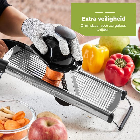 Hand met snijhandschoen snijdt wortel met mandoline. Groenten zoals paprika, peer en appel op de achtergrond.