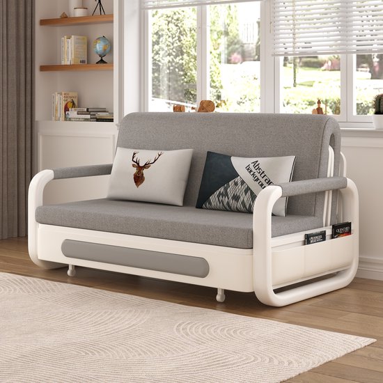 Grijze slaapbank met witte armleuningen, decoratieve kussens en opbergvakken. Geplaatst op een vloerkleed.