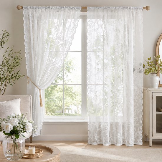 Een woonkamer met een bank, salontafel en een raam bedekt met twee witte bloemenkant vitrages. Een vitrage is vastgebonden met een goudkleurig koord.