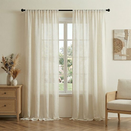 Twee naturelkleurige, lichtdoorlatende gordijnen hangen voor een raam. Een vaas met gedroogde bloemen staat op een houten kastje. Een stoel met rotan details staat ernaast.
