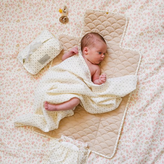 Baby in een luier, gewikkeld in een wit dekentje met beige stippen, liggend op een crèmekleurige aankleedmat met beige patroon. Naast de mat liggen een tissuendoos, slabbetje en twee spenen.