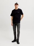 Man met pet, draagt zwart T-shirt met logo, donkergrijze jeans en zwarte schoenen.