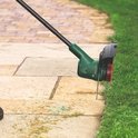 Close-up van de groene kop van een Bosch grastrimmer die langs de rand van een betegeld pad snijdt.