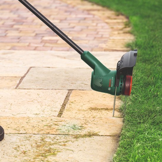 Close-up van de groene kop van een Bosch grastrimmer die langs de rand van een betegeld pad snijdt.