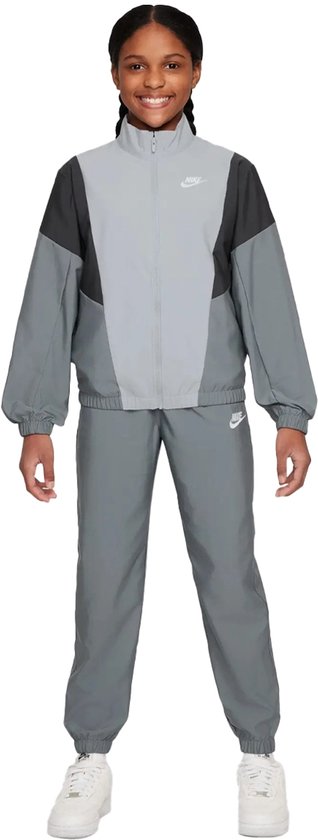 Meisje in een grijs Nike trainingspak met een colorblock design van lichtgrijs, donkergrijs en zwart. Nike logo's op de linkerborst en linkerbroekspijp.