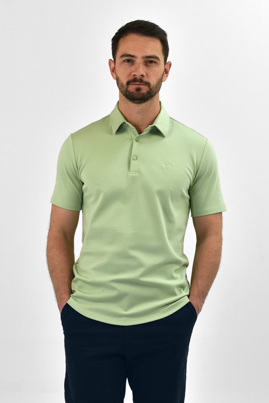 Man in lichtgroene Vercate polo met korte mouwen en donkerblauwe broek, handen in de zakken.