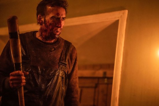 Nicolas Cage, met bloed op zijn gezicht, draagt een bruine trui en overall met een honkbalknuppel.
