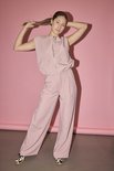 Model in een lichtroze wijde broek en een bijpassend mouwloos topje met rits, staand tegen een roze achtergrond.