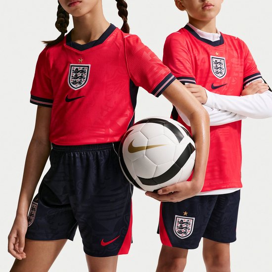Kinderen in rode shirts en marineblauwe shorts, één houdt een voetbal vast.