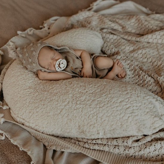 Pasgeboren baby slaapt op een beige voedingskussen, draagt een beige mutsje met pompon.