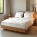 Een opgemaakt bed met een wit dekbed en kussens met beige bladmotieven, bij een raam en houten nachtkastjes.