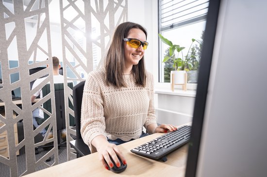 Vrouw met blauwlichtfilterbril met gele glazen werkt op kantoor achter een computer.