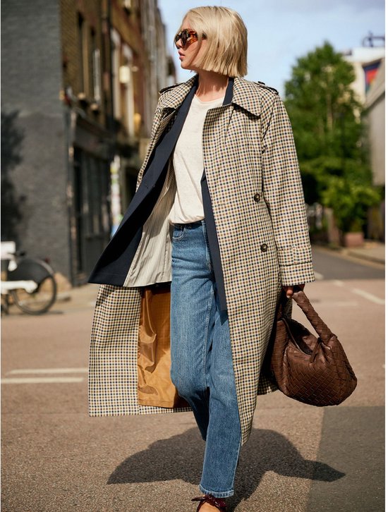 Een vrouw met blond haar, een zonnebril, een geruite trenchcoat, een witte top en een spijkerbroek, lopend door een straat.
