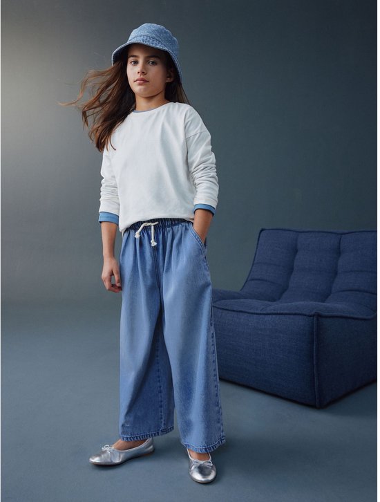 Meisje met bruin haar, staand, draagt wijde, ronde jeans en denim hoed.