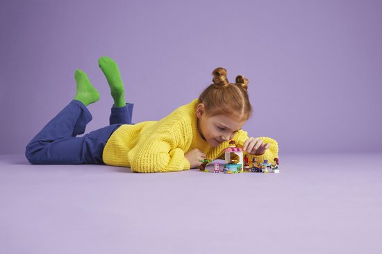 Fille aux couettes rousses et pull jaune jouant avec la Crêperie LEGO Friends sur fond violet.