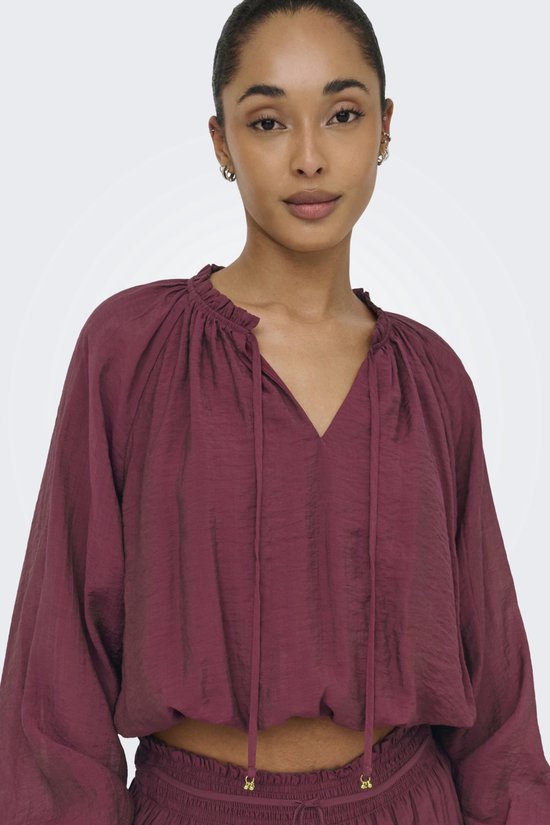 Model draagt een bordeauxrode blouse met strikdetail en lange, wijde mouwen.