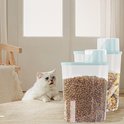 Witte kat likt zijn lippen naast een doorzichtige bak met hondenvoer en een bak met mungbonen.