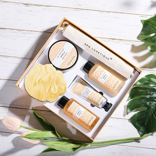 Een geopende geschenkdoos met 'Spa Luxetique Vanilla' badproducten, een badspons en bladeren op een witte houten achtergrond.