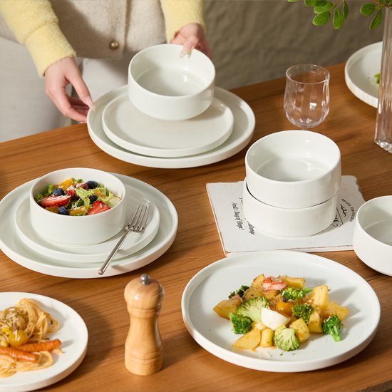 Een vrouw legt witte borden en kommen op een houten tafel, waar ook salade en pasta op staan.